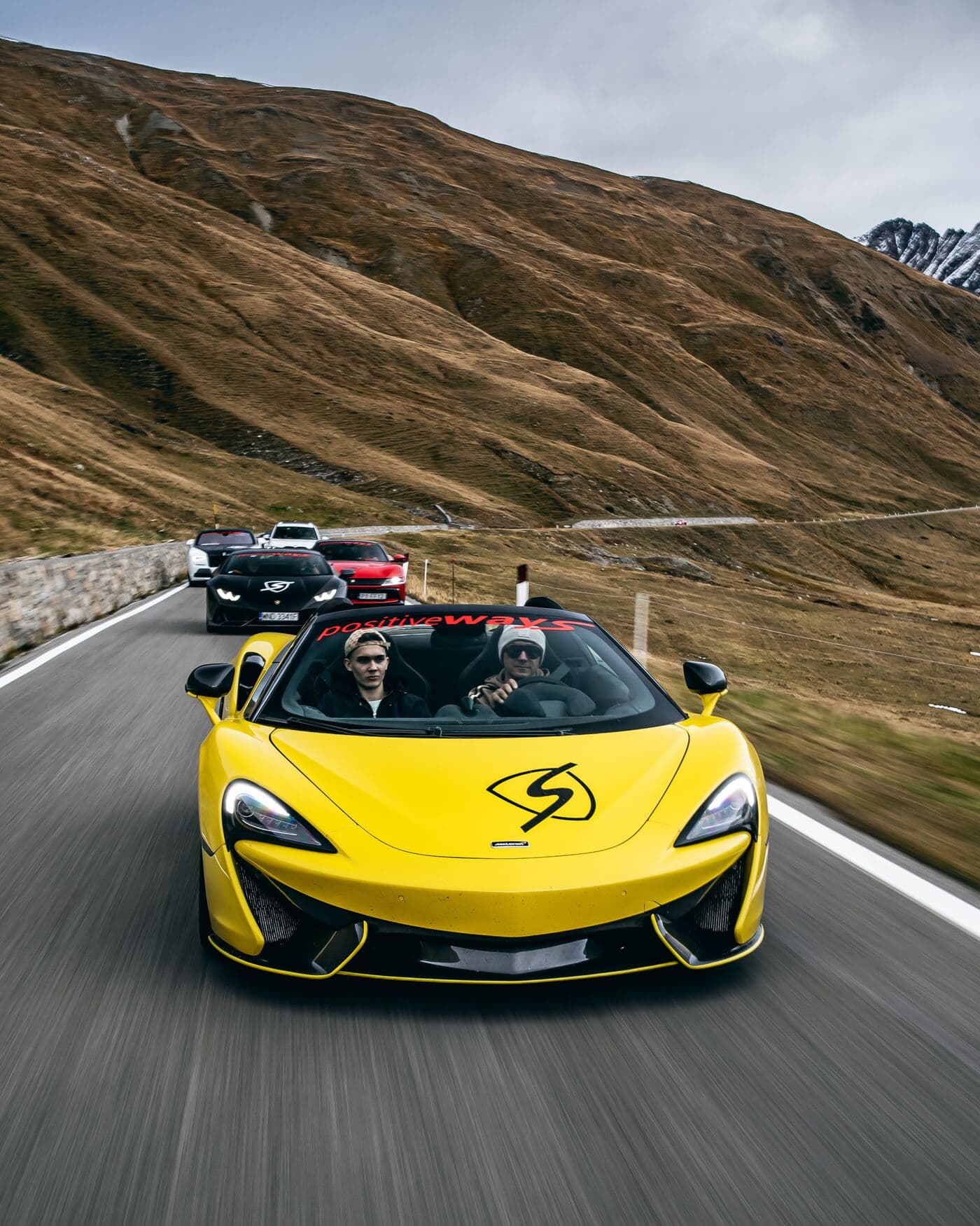 Żółty McLaren prowadzi kolumnę aut na alpejskiej drodze